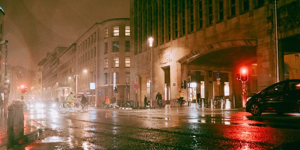 A wet city street at dusk with parked cars and traffic lights